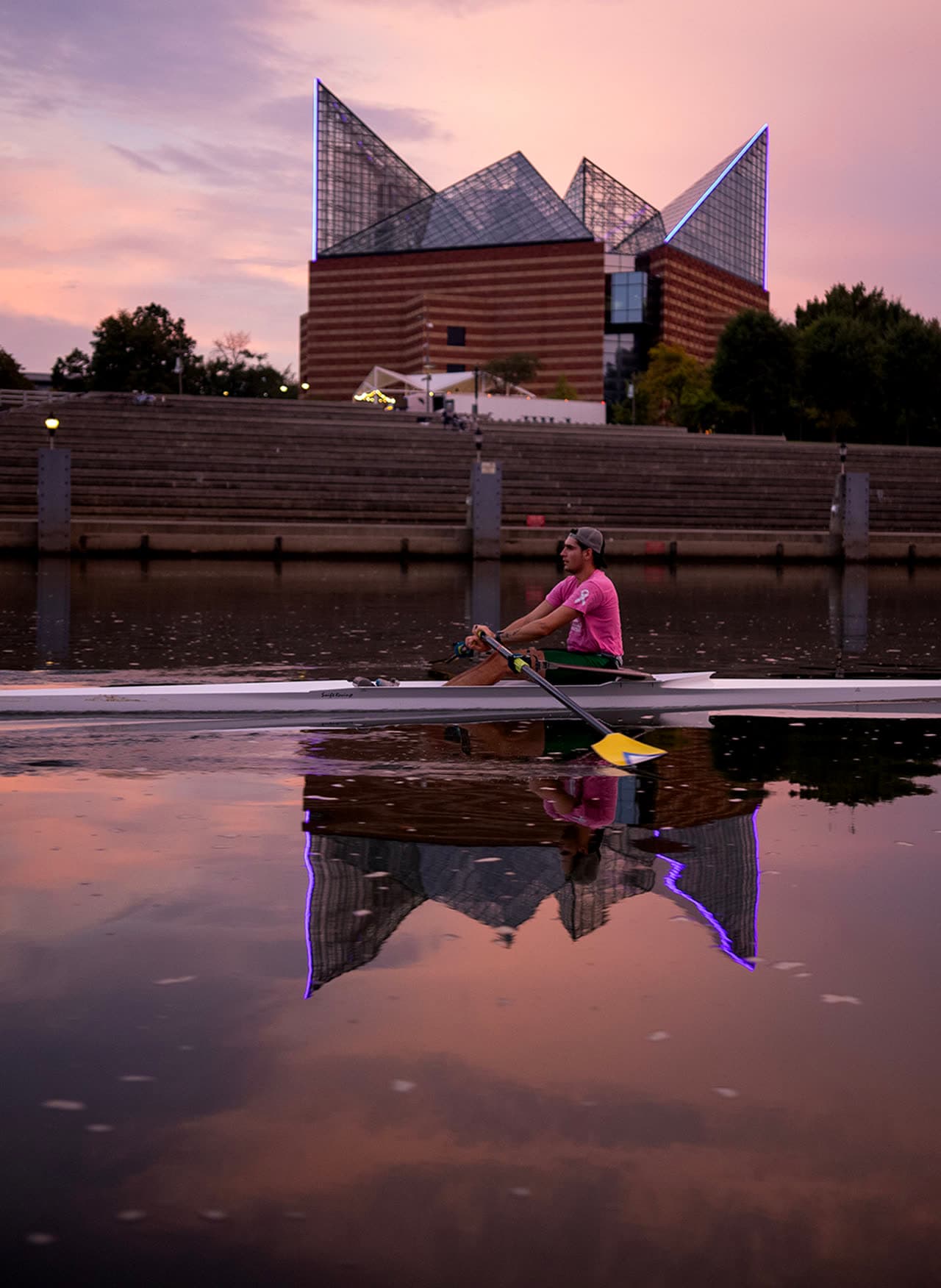 Abraham Mako rowing a single past the Tennessee Aquarium at sunset, the glass pyramid reflected in the river