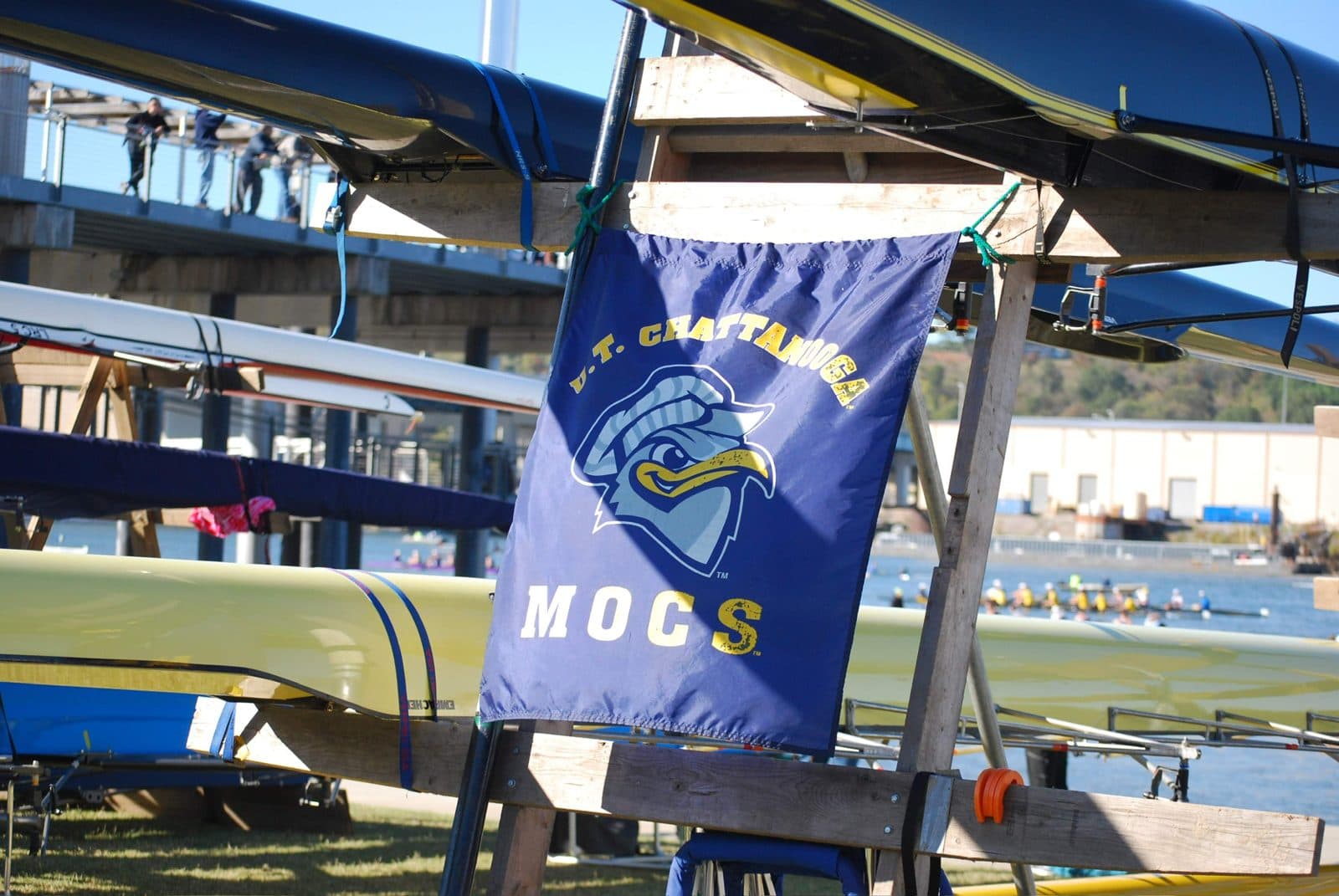 A U.T. Chattanooga Mocs banner hung on UTC's boat racks at the Head of the Hooch, with crews racing on the Tennessee River behind.