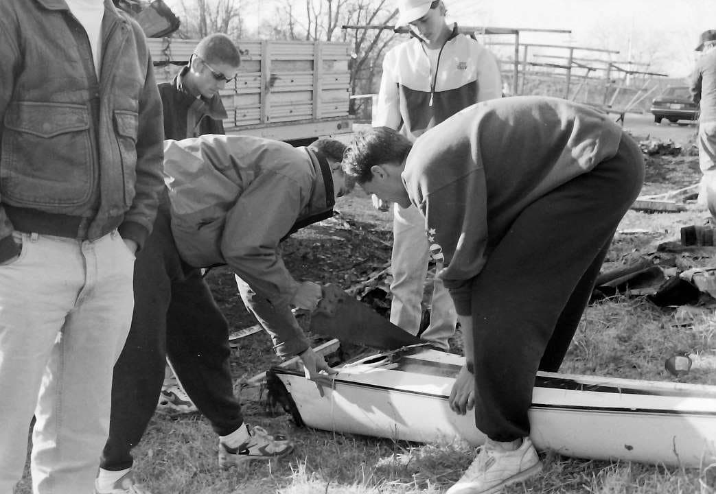 A group of UTC Rowing athletes lifting a salvaged shell piece together in a winter field.