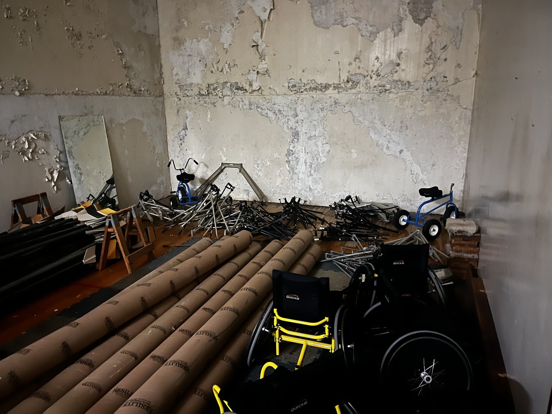 The Maclellan Gym indoor training room — water-damaged walls, rowing equipment and oars stacked against the walls, wheelchairs from the HHP Sports Lab in the foreground