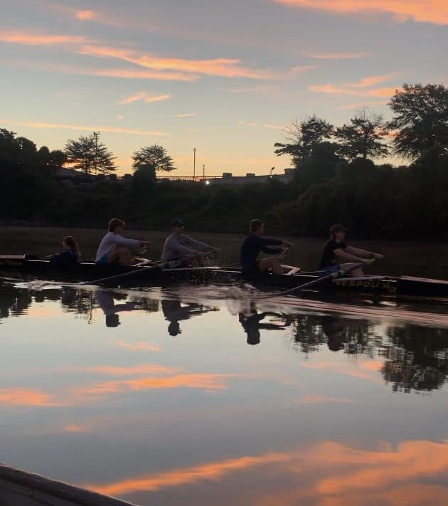 A UTC eight rowing on glassy water at sunset, the crew silhouetted against an orange sky.