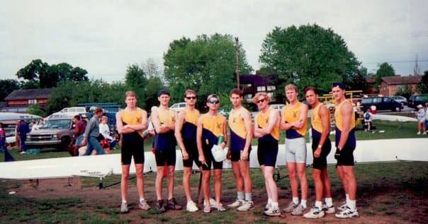 Nine UTC men in yellow/blue jerseys lined up at a regatta venue, several with medals around necks